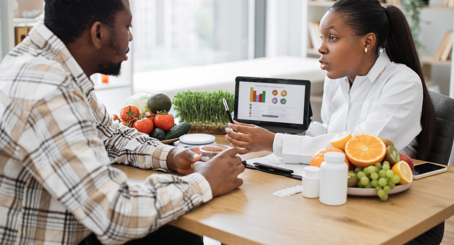 A nutritionist in a white lab coat is sitting at a desk and speaking with a male client wearing a plaid shirt. They are discussing a healthy diet, with a tablet displaying a colorful nutrition chart between them. 