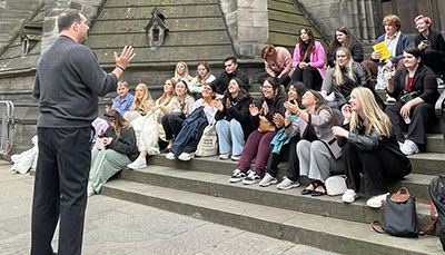 Large group of people sitting on the steps of an foreign building listening to someone standing in front of them.