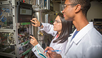 Two students in lab coats and safety glasses working on a device.
