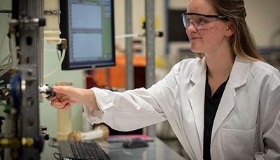 Student in a lab coat and safety glasses interacting with a device.