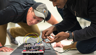 Two students on the ground fixing a machine with wheels and wires. 
