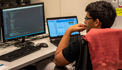 Student sitting in front of a laptop and computer monitor with code on the screen.