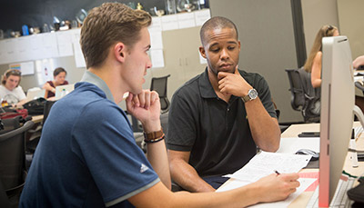 Two students sitting in front of a computer talking with each other.