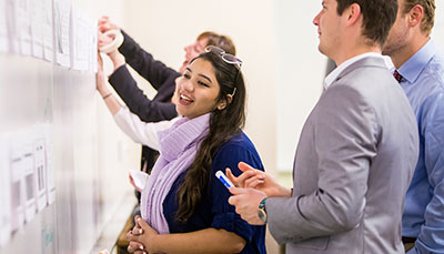 Several students in a classroom standing at a whiteboard looking through papers tapped onto it.