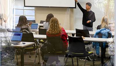 Looking though a glass window into a small class of students with a professor standing in the front.